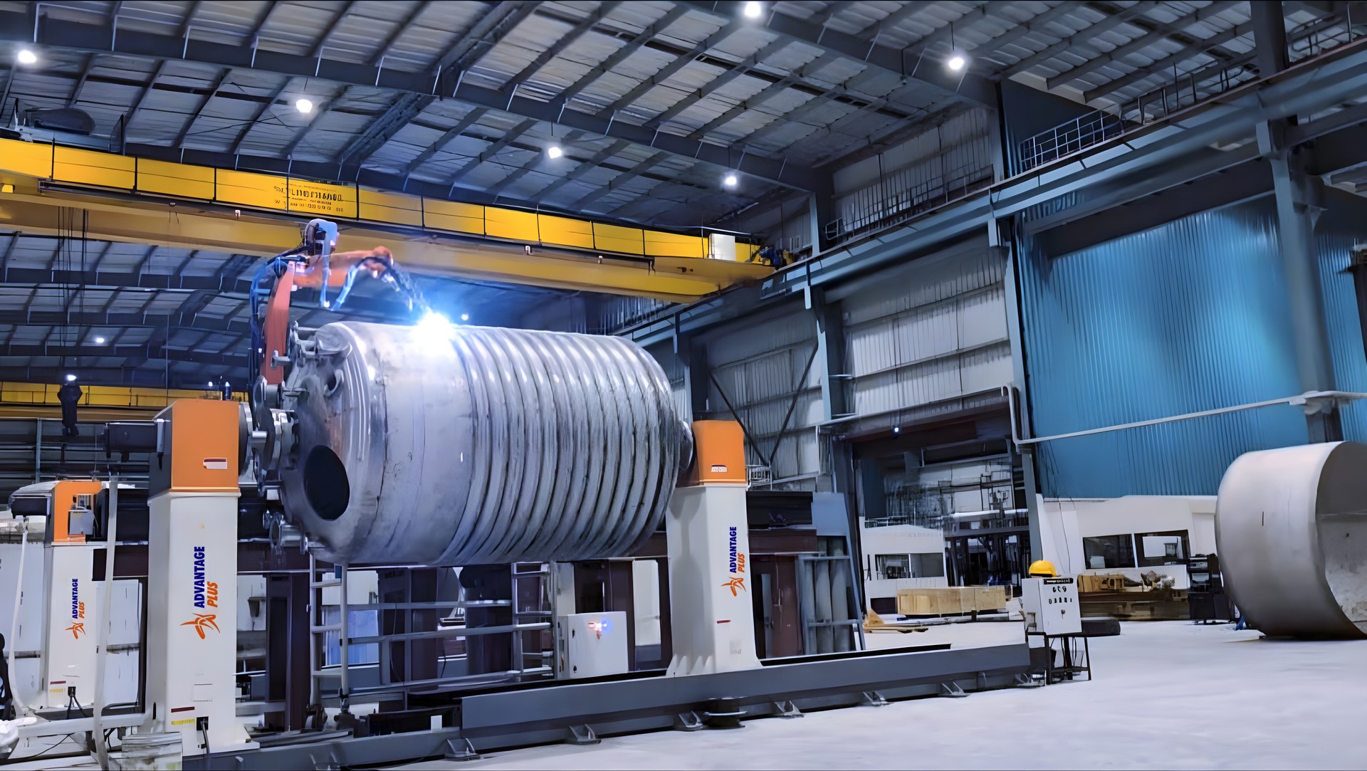 Advantage Plus robotic welding cell: a large limpet-coil vessel held between two positioner columns branded Advantage Plus while an orange welding robot welds the coil, in a large fabrication shop